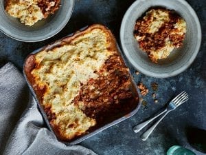 Oven dish of Porcini Shepherd's Pie with two dished up plates on a grey table with accompanying forks