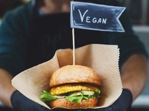 Man holding a burger labelled 'vegan'|100% Vegan chalk sign with an arrow pointing to the left||Restaurant boards showing vegan options including Greggs